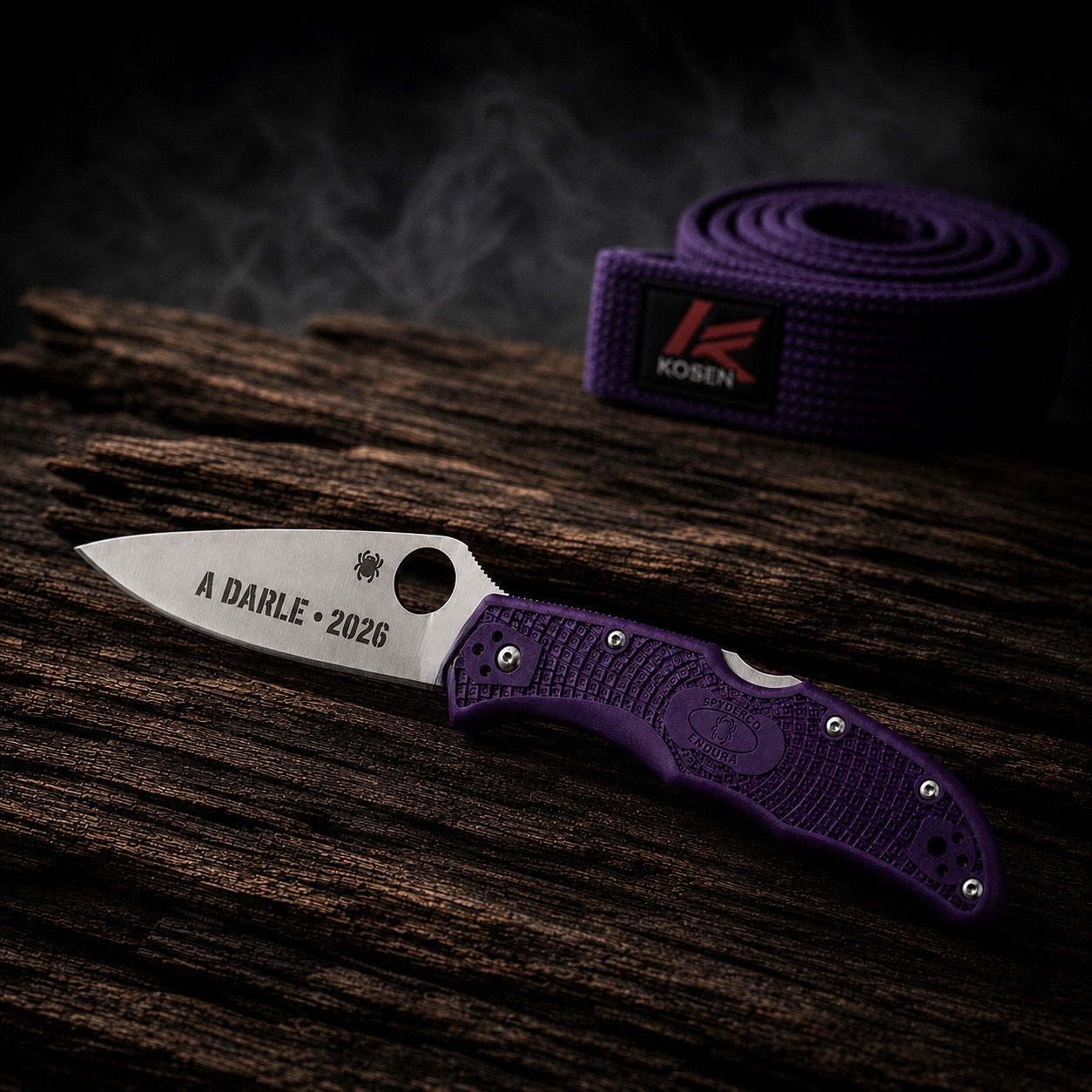 Purple Brazilian jiu-jitsu belt beside a purple-handled Spyderco folding knife engraved “A DARLE • 2026.”